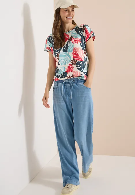 Vrouw in bloemenprint shirt, wijde spijkerbroek en sneakers voor neutrale achtergrond.