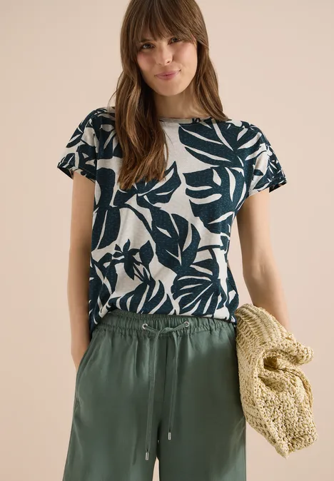 Vrouw met wit t-shirt met bladprint, losse groene broek en grote strozak.