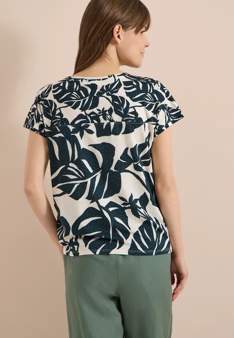 Achteraanzicht van een vrouw met een wit T-shirt met donker bladpatroon en een groene broek.