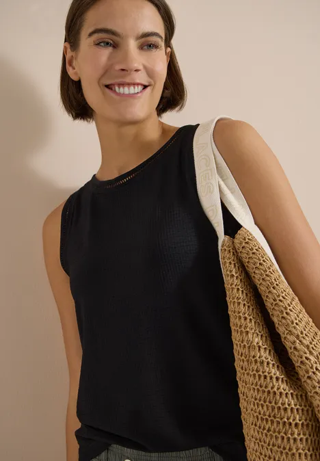 Frau in schwarzem ärmellosem Top mit großem geflochtenem Strohtasche vor neutralem Hintergrund.