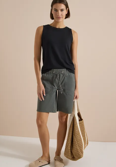 Frau in ärmellosem schwarzen Top, grauen Shorts und Sandalen, trägt eine geflochtene Strandtasche.