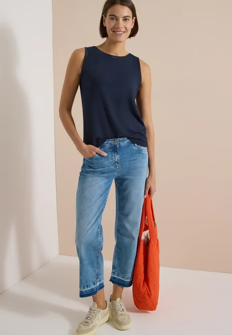 Gut angezogene Frau mit ärmellosem Marineblau-Top, hellblauen Jeans, Sneakern und roter Stofftasche.