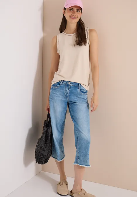 Top met boothals en kanten details pearl beige Frau in beigem ärmellosem Top, hellblauen Capri-Jeans und Sandalen trägt eine schwarze Tasche vor einer Wand.