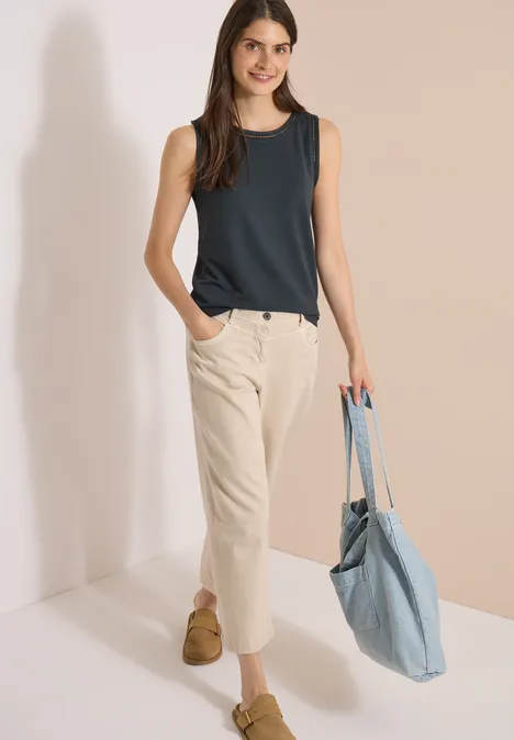 Top met boothals en kanten details palm breeze green Frau trägt dunkles ärmelloses Top, helle Hose und blaue Stofftasche.