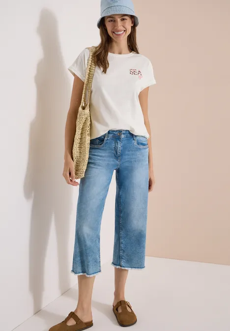 Shirt met ronde hals en borduursel vanilla white Vrouw met wit T-shirt, brede cropped jeans, pet, bruine instappers en strozak tegen beige-witte achtergrond.