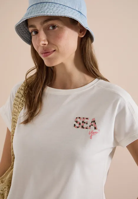 Shirt met ronde hals en borduursel vanilla white Vrouw met lichtblauwe vissershoed, wit t-shirt met “SEA you”-print en strozak.
