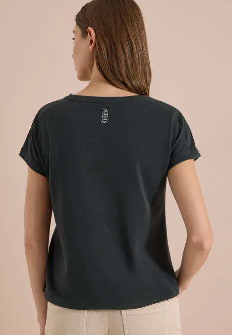 Shirt met ronde hals en borduursel palm breeze green Een vrouw van achteren draagt een zwart T-shirt met omgeslagen mouwen en een beige broek voor een neutrale achtergrond.