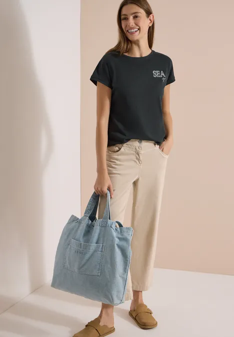 Shirt met ronde hals en borduursel palm breeze green Vrouw draagt zwart T-shirt, beige broek en bruine loafers en houdt een lichtblauwe spijkertas vast.
