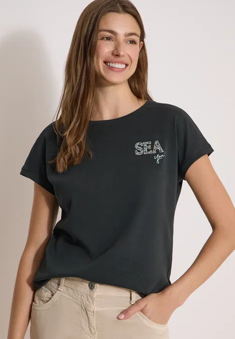 Shirt met ronde hals en borduursel palm breeze green Vrouw in een zwart 'SEA'-T-shirt en beige broek, nonchalant staand met één hand in de zak.