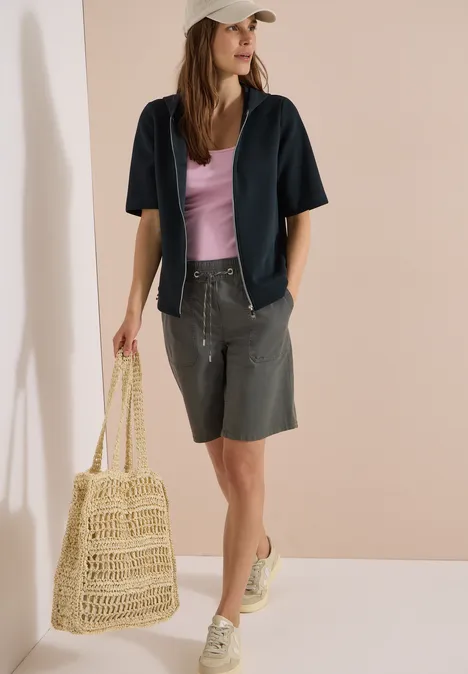 Kapuzenjacke mit seitlichen Tunnelzügen palm breeze green Frau trägt graue Bermudashorts, helles Top, dunkelblaues Hemd, Mütze, Strohtasche und Sneaker.