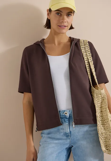Person trägt kurzes braunes Kapuzenshirt, weißes Top, blaue Jeans und Basttasche.