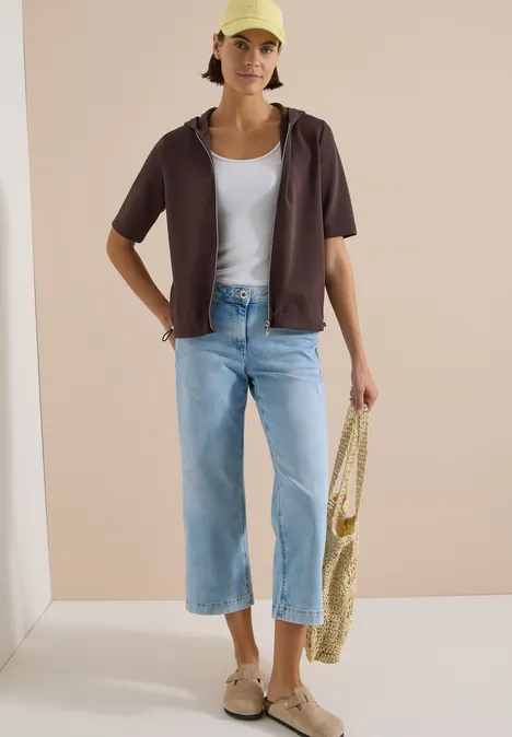 Frau mit weißem Top, kurzarm Hemd, weitem Jeans, Sandalen und Basttasche in minimalistischem setting.