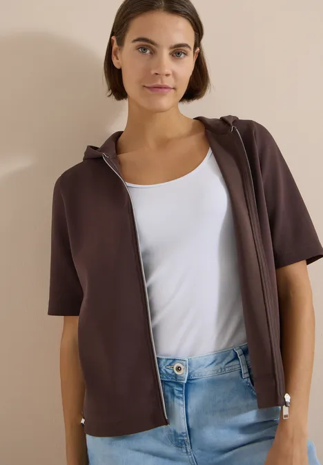 Person trägt braunen kurzärmligen Kapuzen-Sweatshirt mit Reißverschluss, weißen Tanktop und Jeans.