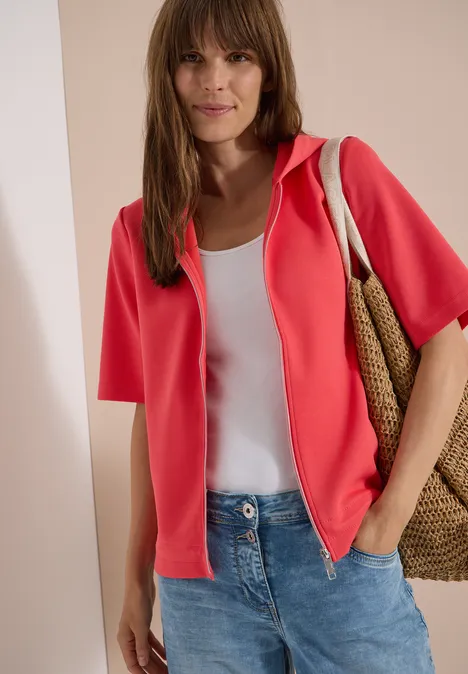 Kapuzenjacke mit seitlichen Tunnelzügen coral Frau trägt rote Kurzarm-Jacke, weißes Top, Jeans und Korbtasche vor neutralem Hintergrund.