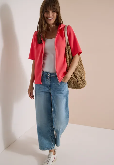 Kapuzenjacke mit seitlichen Tunnelzügen coral Frau trägt weißes Top, pinke Jacke, weite Jeans, weiße Sneaker und Korbtasche vor hellen Wänden.