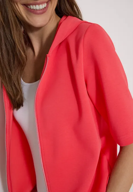 Kapuzenjacke mit seitlichen Tunnelzügen coral Nahaufnahme einer lächelnden Frau in korallenrotem, kurzärmeligem Hoodie und weißem Top