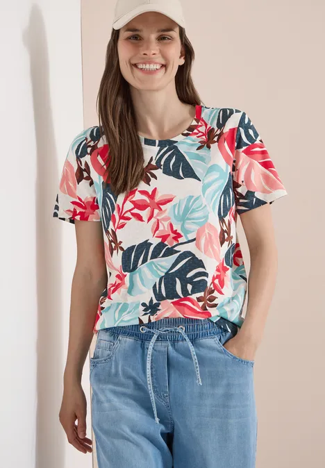 T-Shirt mit Rundhals und Blätterprint light alabaster white Frau trägt kurzärmlige Bluse mit rotem Blumen- und blauem Blattmuster und hellblauen Jeans.