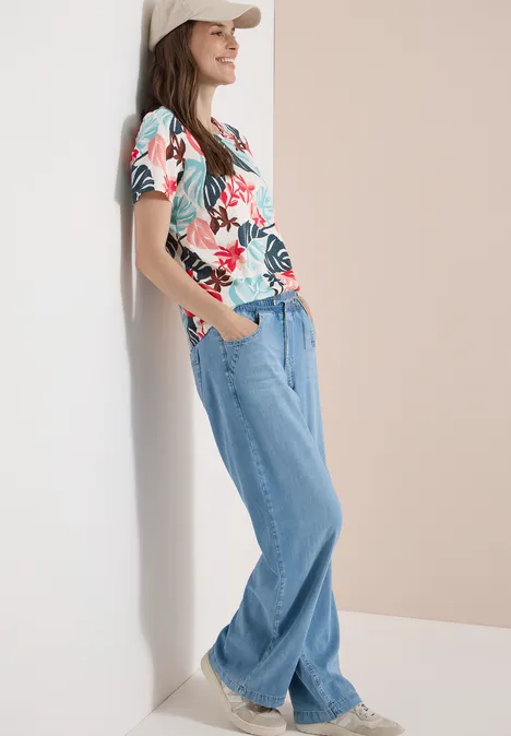 T-Shirt mit Rundhals und Blätterprint light alabaster white Eine Person lehnt entspannt an einer Wand, trägt ein buntes Blumen-T-Shirt, weite Jeans und weiße Sneaker.
