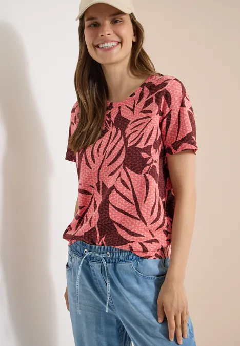T-Shirt mit Rundhals und Blätterprint coral Frau im kurzärmligen, korallrosa Shirt mit tropischen Blattmotiven und hellblauen Jeans-Shorts.