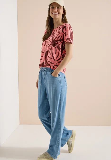 T-Shirt mit Rundhals und Blätterprint coral Frau mit pinkem Blattmuster-T-Shirt und lockeren Jeans vor neutralem Hintergrund.
