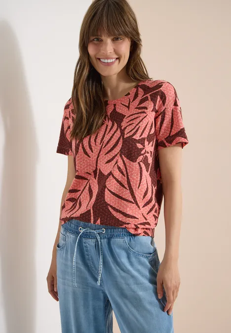 T-Shirt mit Rundhals und Blätterprint coral Person in rotem Kurzarmshirt mit Blattmuster und blauer Kordel-Jeans, stehend vor hellem Hintergrund.