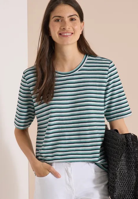 T-Shirt mit Struktur und Streifen palm breeze green Frau mit gestreiftem Kurzarmshirt, weißer Hose und schwarzer Tasche vor beigem Hintergrund.