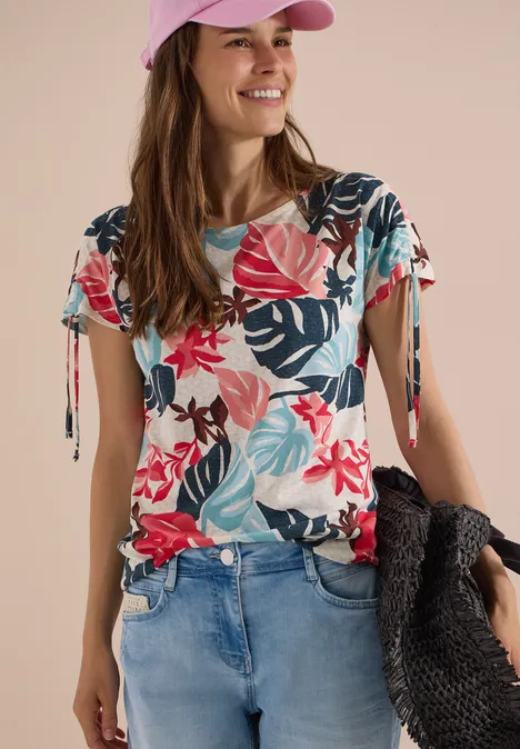Frau in Jeans, buntem Blumen-Shirt und rosa Cap hält einen schwarzen Korb.