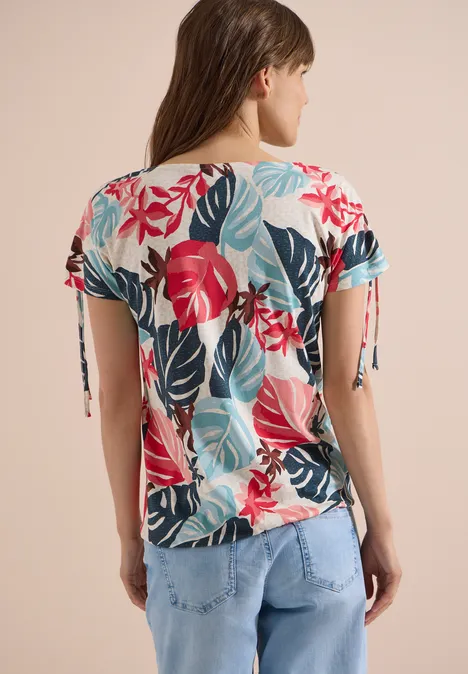 Frau von hinten in blumigem, kurzärmeligem Shirt mit tropischem Blattmuster und hellen Jeans.