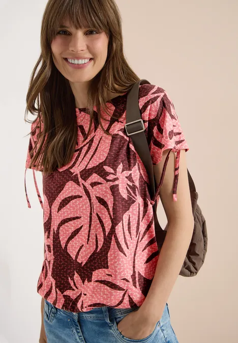 Vrouw met roze bladpatroon-shirt, spijkerbroek en rugzak voor lichte achtergrond.