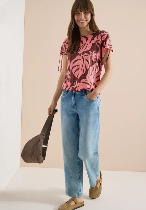 Vrouw in roze tropische blouse, lichte jeans, gevlochten sandalen en een bruin tasje.