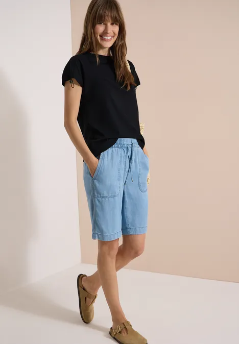 T-Shirt mit Raffung in Unifarbe Black Frau in schwarzem T-Shirt, hellblauen Jeans-Shorts und braunen Slippern vor schlichtem, hellem Hintergrund.