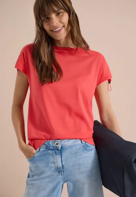 Frau trägt rotes T-Shirt, blaue Jeans und hält eine schwarze Jacke.