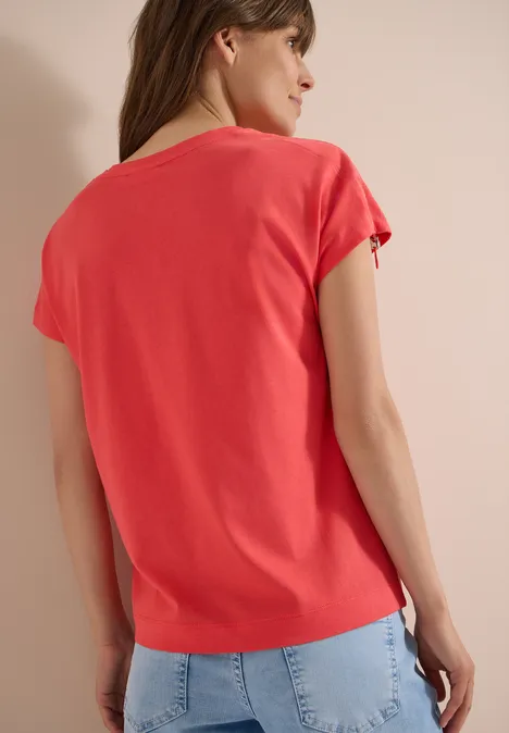 Frau von hinten in korallrotem T-Shirt und hellblauer Jeans vor neutralem Hintergrund.