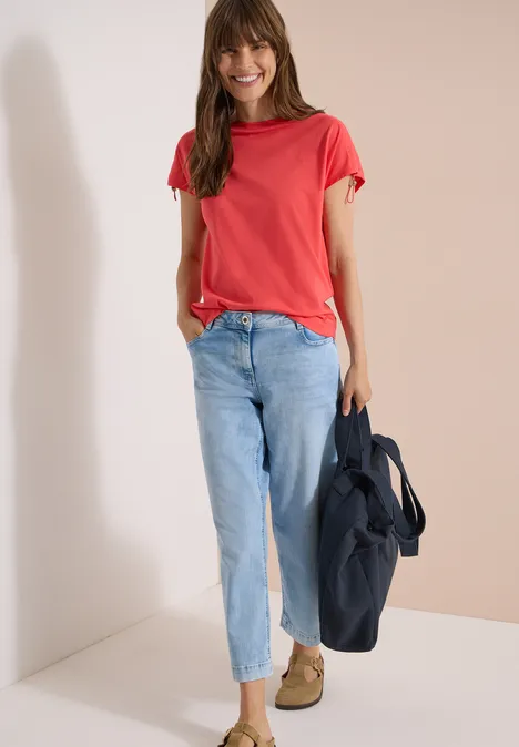 Eine Person steht vor weißem Hintergrund, trägt rotes T-Shirt, helle Jeans und hält eine dunkle Tasche.