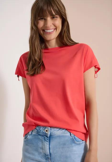 Frau in korallenrotem T-Shirt mit kurzen Ärmeln und Jeans vor hellem Hintergrund.