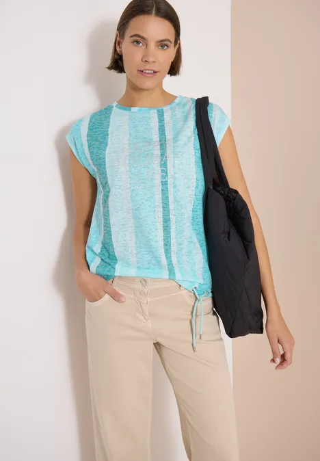 T-Shirt in Ausbrenner-Optik crystal mint Frau in türkis-weiß gestreiftem Top, beiger Hose und schwarzer Tragetasche vor neutralem Hintergrund.