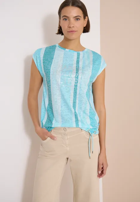 T-Shirt in Ausbrenner-Optik crystal mint Frau in blau-weiß gestreiftem Top und beiger Hose vor hellem Hintergrund.