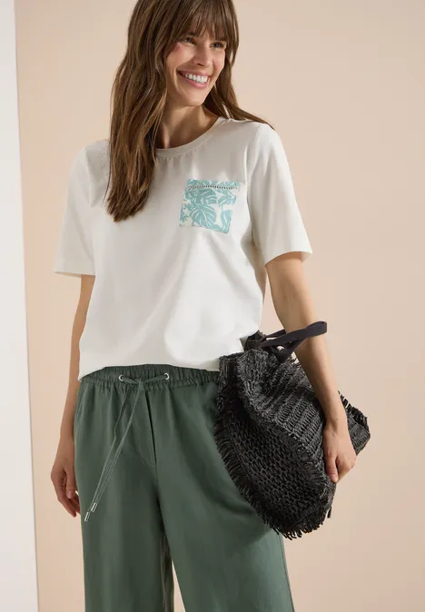 T-Shirt mit Rundhals und Brusttasche vanilla white Frau trägt weißes T-Shirt mit türkisem Brusttaschen-Print, weite grüne Hose und großen schwarzen Netzbeutel.