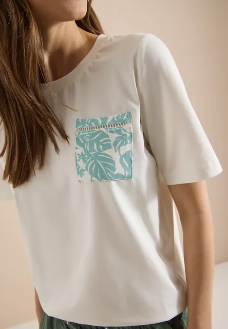 T-Shirt mit Rundhals und Brusttasche vanilla white Weißes T-Shirt mit türkisem tropischem Blattmuster auf der Brusttasche, getragen von einer Frau.