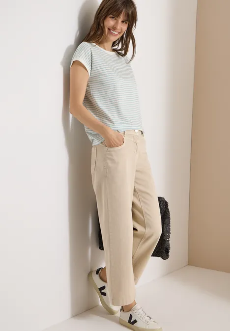 T-Shirt mit Boat Neck und Streifen crystal mint Frau lehnt entspannt an einer Wand und trägt ein gestreiftes T-Shirt, beige Hose und weiße Sneaker.