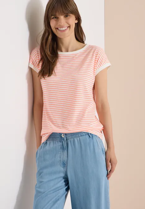 T-Shirt mit Boat Neck und Streifen coral Frau im gestreiften T-Shirt und leichter Jeanshose lehnt an hellem Hintergrund.