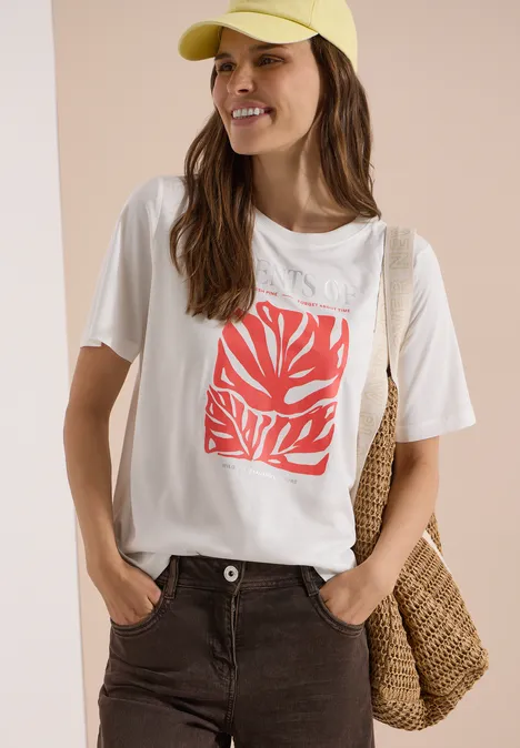 Frau mit gelber Mütze, weißem T-Shirt mit rotem Blattmotiv, braunen Hosen und Korbtasche vor hellem Hintergrund.