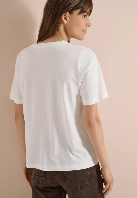 Frau steht mit dem Rücken zur Kamera und trägt ein schlichtes weißes T-Shirt und braune Hose vor beigem Hintergrund.
