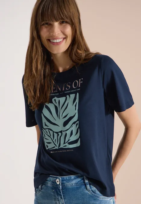 T-Shirt mit Rundhals und Frontprint universal blue Frau in dunkelblauem T-Shirt mit grünem Blattmuster und blauen Jeans.