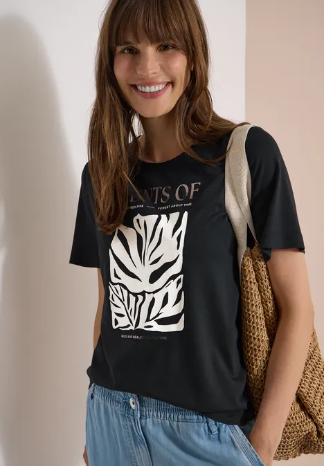 T-Shirt mit Rundhals und Frontprint palm breeze green Frau vor neutralem Hintergrund, trägt schwarzes T-Shirt mit weißem Zebramuster, blaue Jeans, Strohtasche.