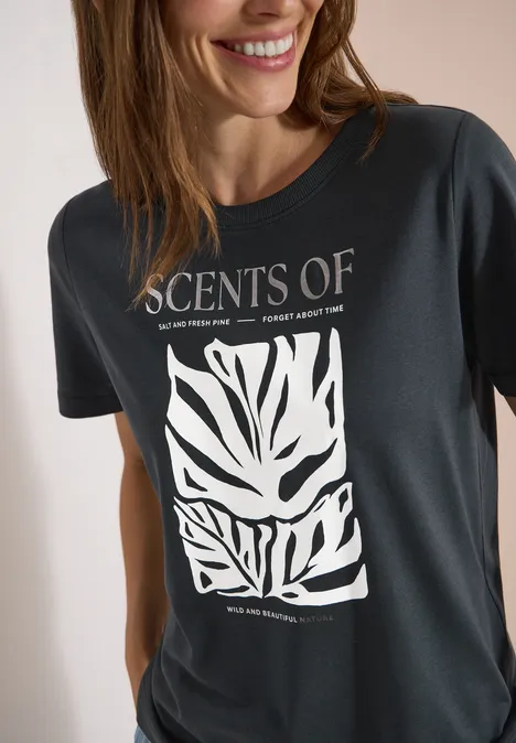 T-Shirt mit Rundhals und Frontprint palm breeze green Frau in dunklem T-Shirt mit Aufdruck 'SCENTS OF' und weißem, abstraktem Blattmuster.