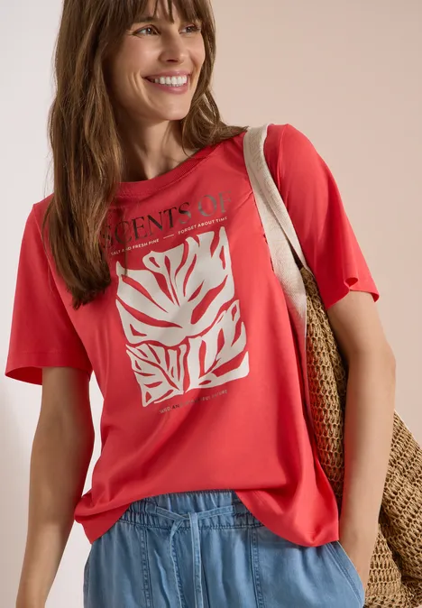 T-Shirt mit Rundhals und Frontprint coral Frau trägt rotes T-Shirt mit weißem Blatt-Print, hellblaue Jeans und Basttasche über der Schulter.