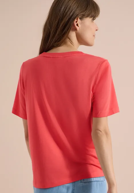 T-Shirt mit Rundhals und Frontprint coral Frau steht mit dem Rücken zur Kamera, trägt rotes T-Shirt und Jeans vor hellem Hintergrund.