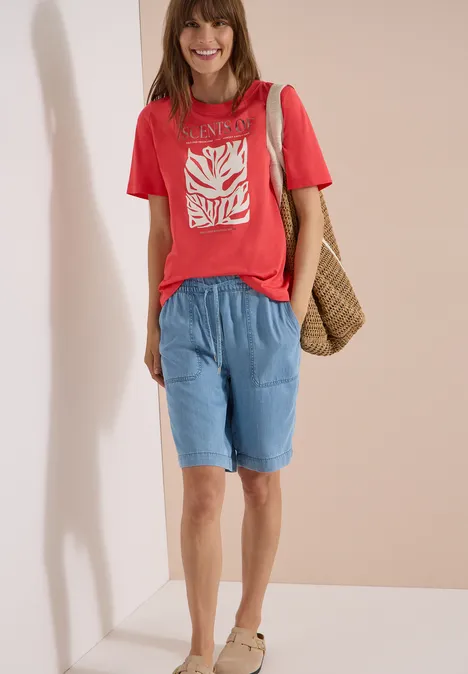 T-Shirt mit Rundhals und Frontprint coral Eine Frau trägt rotes T-Shirt mit weißem Motiv, lockere hellblaue Shorts und eine Korbtasche.