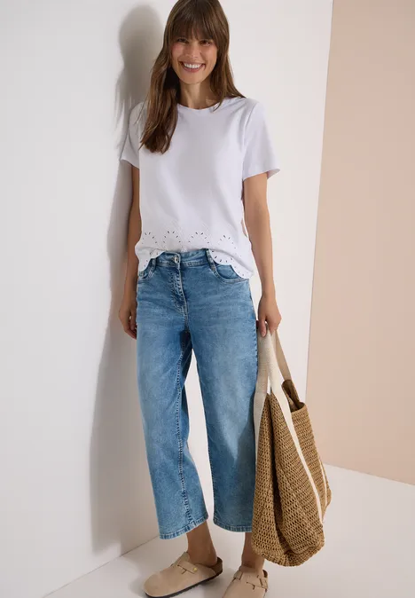 Frau in weißem T-Shirt mit Spitzensaum, weitem Jeans, Slip-on-Schuhen und Strohtasche vor hellem Hintergrund.